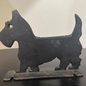May include: A black cast iron doorstop shaped like a Scottish Terrier dog. The silhouette features a pointed ear, a short tail, and a rectangular base. The metal surface shows signs of wear and minor rust along the edges and the base.