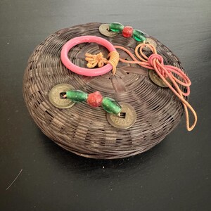 May include: A round, woven basket with a pink ring and decorative coins. The basket is brown and has a textured surface. Green and red beads are strung on a cord with the coins.