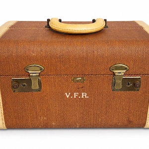May include: A rectangular brown suitcase with a textured fabric finish and cream-colored trim. It features a top handle, two brass-toned metal latches, and the initials V.F.R. printed in white on the front center. A small logo tag is visible.