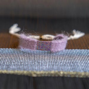 May include: A woven bracelet with a lavender band and a multicoloured centre section. The bracelet has white tassels on each end. The bracelet is displayed on a textured surface with blue and brown tones.