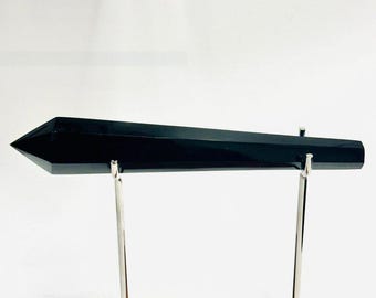 May include: A long, polished black obsidian crystal wand with a pointed tip, held horizontally by two silver-toned metal prongs attached to a rectangular wooden base. The crystal has a smooth, reflective surface and a hexagonal faceted shape.