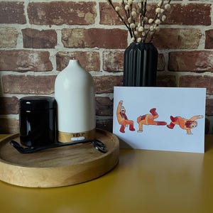 The Simpsons Planet of the Apes print, sat on a on a yellow desk propped up on a black vase with pussy willow branches is in the background. Surrounded with a white oil diffuser, a black candle jar, and a black wick trimmer on a wooden tray. Showing what the print would look like in a home set up.