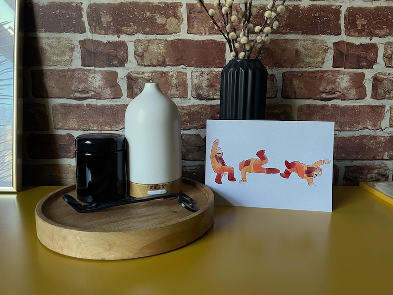 The Simpsons Planet of the Apes print, sat on a on a yellow desk propped up on a black vase with pussy willow branches is in the background. Surrounded with a white oil diffuser, a black candle jar, and a black wick trimmer on a wooden tray. Showing what the print would look like in a home set up.