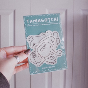 May include: A set of three Tamagotchi embroidery templates on a blue background. The templates are white and feature different Tamagotchi characters. The text "Tamagotchi" and "stitch & stitch embroidery templates" is printed on the card. The text "Fawn & Flora" is printed at the bottom of the card.