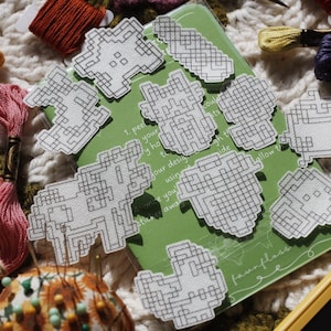Könnte beinhalten: Ein Set mit acht weißen Kreuzstichmustern auf grünem Hintergrund. Die Muster sind in verschiedenen Formen und Größen, darunter Herzen, Sterne und geometrische Designs. Die Muster sind auf einem Blatt Papier mit Anweisungen zum Sticken gedruckt. Das Papier liegt auf einer weißen Oberfläche mit buntem Stickgarn.