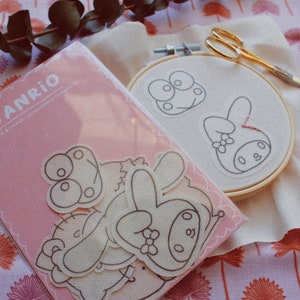 May include: A pink and white embroidery kit featuring Sanrio characters. The kit includes a pre-printed fabric, embroidery floss, and a needle. The characters include a frog and a bunny.