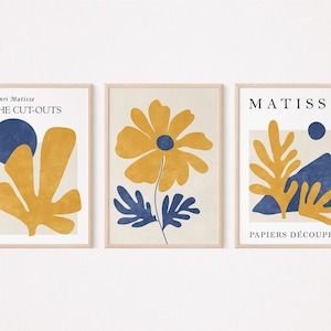 Puede incluir: Tres impresiones enmarcadas con arte abstracto en amarillo y azul. La primera impresión tiene el texto "Henri Matisse THE CUT-OUTS". La segunda impresión presenta una flor amarilla con hojas azules. La tercera impresión tiene el texto "MATISSE PAPIERS DÉCOUPÉS".