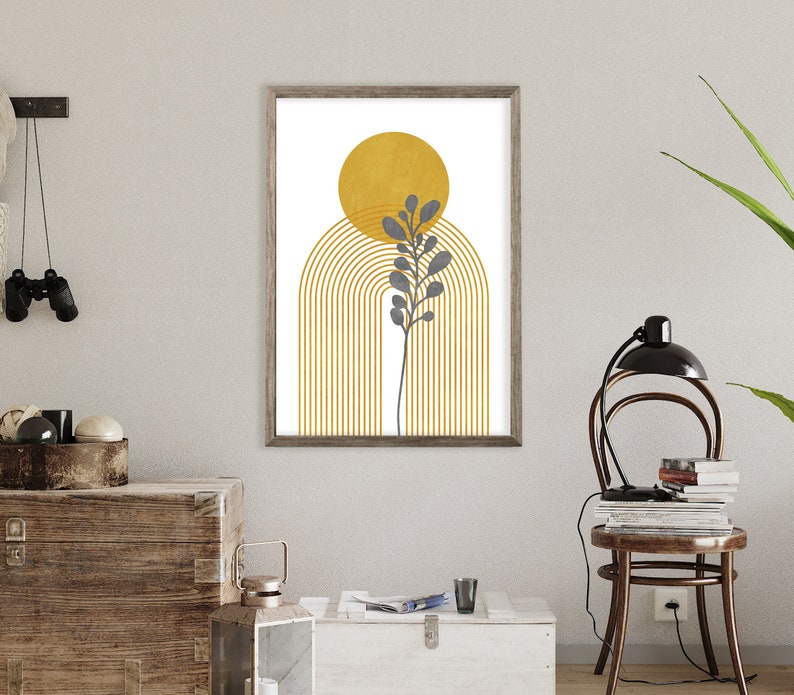May include: A framed print with a minimalist design featuring a yellow sun, a gray plant, and a gold archway. The print has a white background and is framed in a light brown wood frame.