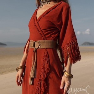 Puede incluir: Un vestido cruzado de color &oacute;xido con flecos en las mangas y la falda. El vestido se ci&ntilde;e a la cintura con un cintur&oacute;n marr&oacute;n con una hebilla circular grande. Brazaletes dorados y un collar gargantilla completan el look bohemio.