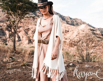 Könnte beinhalten: Eine Frau trägt ein weißes Häkel-Kimono mit Fransen, ein braunes ärmelloses Oberteil und einen breiten braunen Gürtel. Sie trägt einen schwarzen Cowboyhut und hat lange braune Haare. Sie steht auf einem Feld mit einem Berg im Hintergrund. Das Wort "kayamé" ist in Kursivschrift auf dem Boden geschrieben.