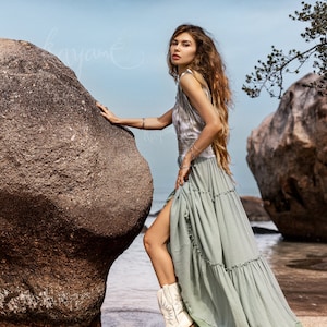 Puede incluir: Una mujer con un vestido maxi verde claro con un top de tie-dye y botas vaqueras blancas posa en una playa con grandes rocas en el fondo.