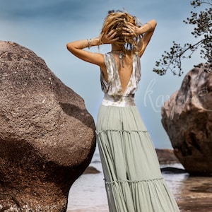 Puede incluir: Una mujer con un vestido maxi verde claro con un top de tie-dye blanco y verde. El vestido tiene la espalda baja y una falda de capas. Est&aacute; de pie en una playa con grandes rocas detr&aacute;s de ella.