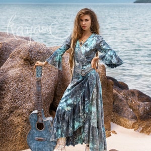 May include: A woman wearing a blue and green tie-dye wrap dress with a silver belt and white cowboy boots. She is standing on a beach with rocks and a guitar beside her.