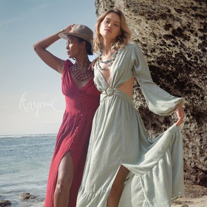 Puede incluir: Dos mujeres con vestidos largos de pie en una playa de arena. Una mujer lleva un vestido rojo con una abertura en el lateral y un sombrero marr&oacute;n. La otra mujer lleva un vestido verde claro con la espalda descubierta y un cintur&oacute;n dorado. Ambas mujeres llevan collares.