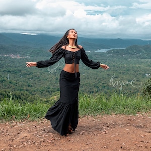 May include: A woman wearing a black off-the-shoulder crop top with a lace-up front and a black maxi skirt. She is standing on a hilltop with a view of a lush green forest and mountains in the distance. The sky is cloudy and the woman is looking up at the sky.