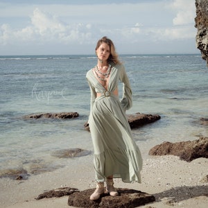 Puede incluir: Una mujer con un vestido maxi verde claro con un dise&ntilde;o de corte y un cintur&oacute;n. Est&aacute; de pie sobre una roca en una playa con el oc&eacute;ano de fondo.