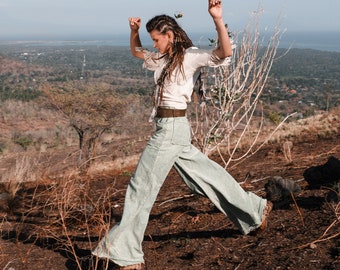 May include: A woman with long brown hair wearing a white shirt and light blue pants is jumping in the air with her arms raised. She is standing on a hill with dry grass and bushes. The sky is blue and there is a distant view of a town.