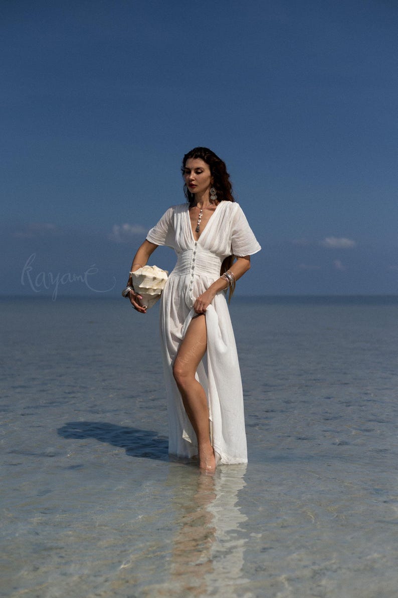 May include: A white, flowing dress with a V-neck and button details. The dress has short sleeves and a high slit. A large seashell is held in the hand. The background is a clear blue sky and ocean.