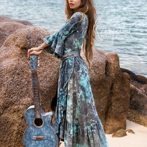 May include: A woman in a blue and white tie-dye dress stands on a sandy beach with a blue acoustic guitar. She is wearing a brown leather belt and white cowboy boots.