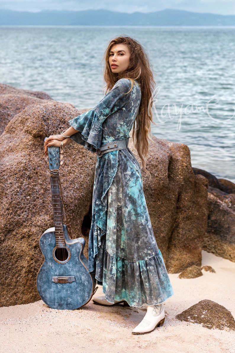 May include: A woman in a blue and white tie-dye dress stands on a sandy beach with a blue acoustic guitar. She is wearing a brown leather belt and white cowboy boots.