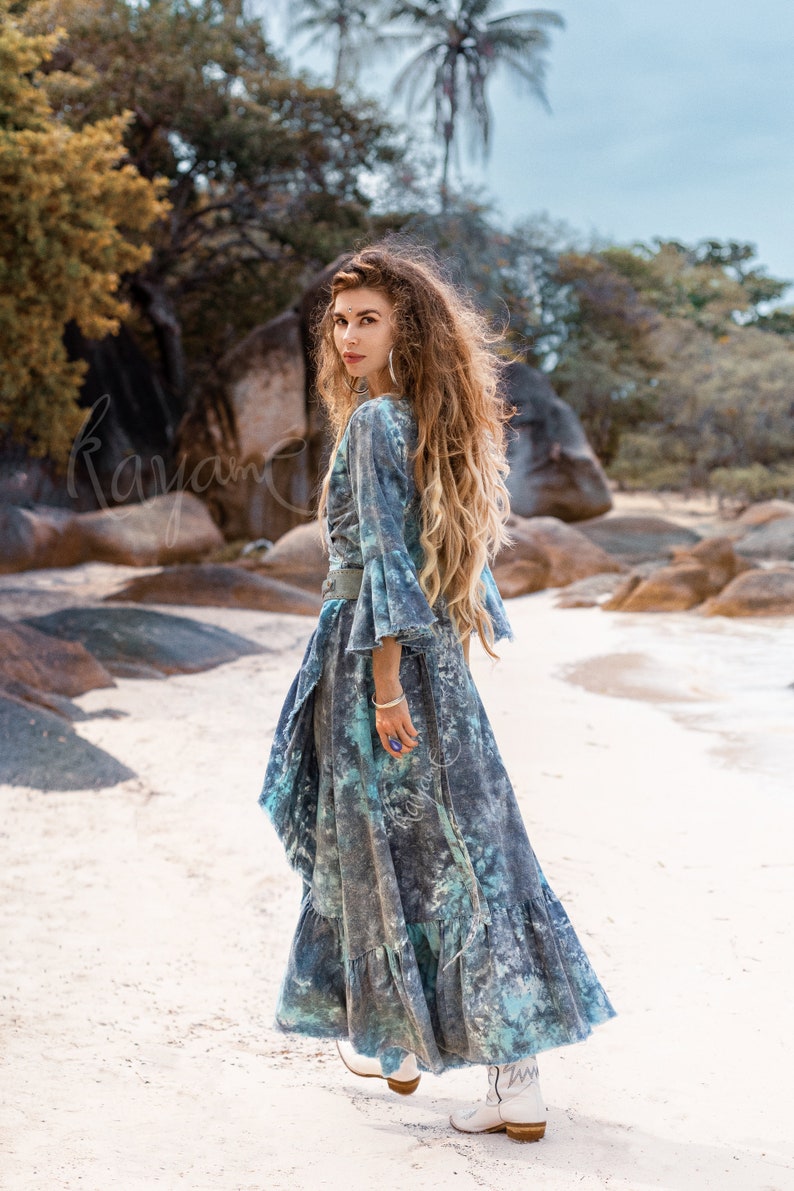 May include: A woman wearing a long, flowing blue and white tie-dye dress with a ruffled hem. She is standing on a sandy beach with rocks and trees in the background. She is wearing white cowboy boots.