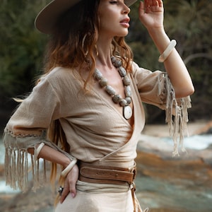 Puede incluir: Una mujer con un vestido cruzado beige, sombrero de ala ancha y cintur&oacute;n de cuero marr&oacute;n. El vestido tiene flecos en las mangas. Tambi&eacute;n lleva un collar de cuentas grande y pulseras. La imagen est&aacute; tomada al aire libre.