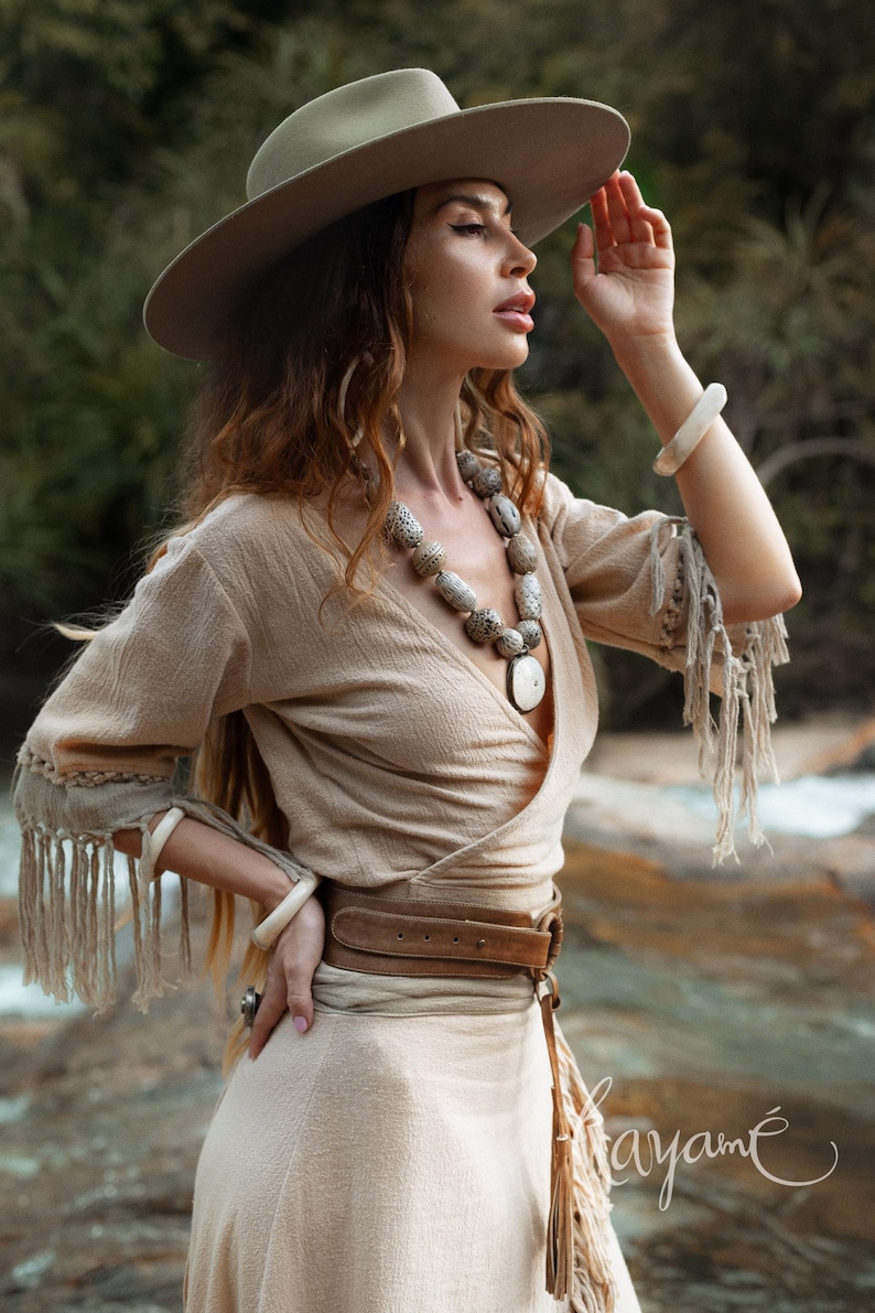 Puede incluir: Una mujer con un vestido cruzado beige, sombrero de ala ancha y cintur&oacute;n de cuero marr&oacute;n. El vestido tiene flecos en las mangas. Tambi&eacute;n lleva un collar de cuentas grande y pulseras. La imagen est&aacute; tomada al aire libre.