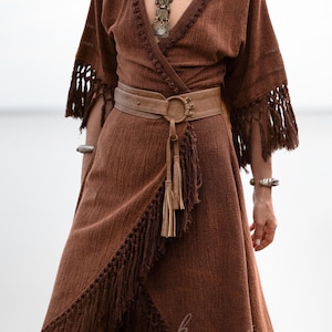 Puede incluir: Vestido cruzado marr&oacute;n con flecos en las mangas y el dobladillo. El vestido se ci&ntilde;e a la cintura con un cintur&oacute;n marr&oacute;n claro con una hebilla circular grande y borlas. Un collar y pulseras plateadas completan el look bohemio.