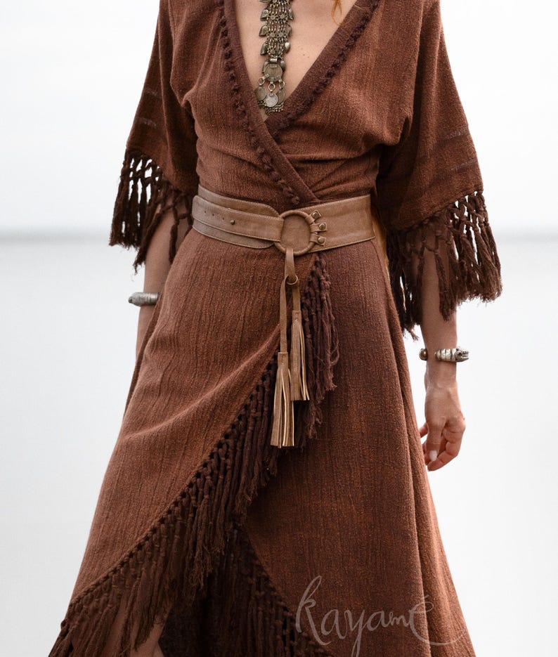 Puede incluir: Vestido cruzado marr&oacute;n con flecos en las mangas y el dobladillo. El vestido se ci&ntilde;e a la cintura con un cintur&oacute;n marr&oacute;n claro con una hebilla circular grande y borlas. Un collar y pulseras plateadas completan el look bohemio.