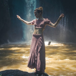May include: A woman wearing a pink skirt and top stands in front of a waterfall. She is facing the waterfall and has her arms outstretched.
