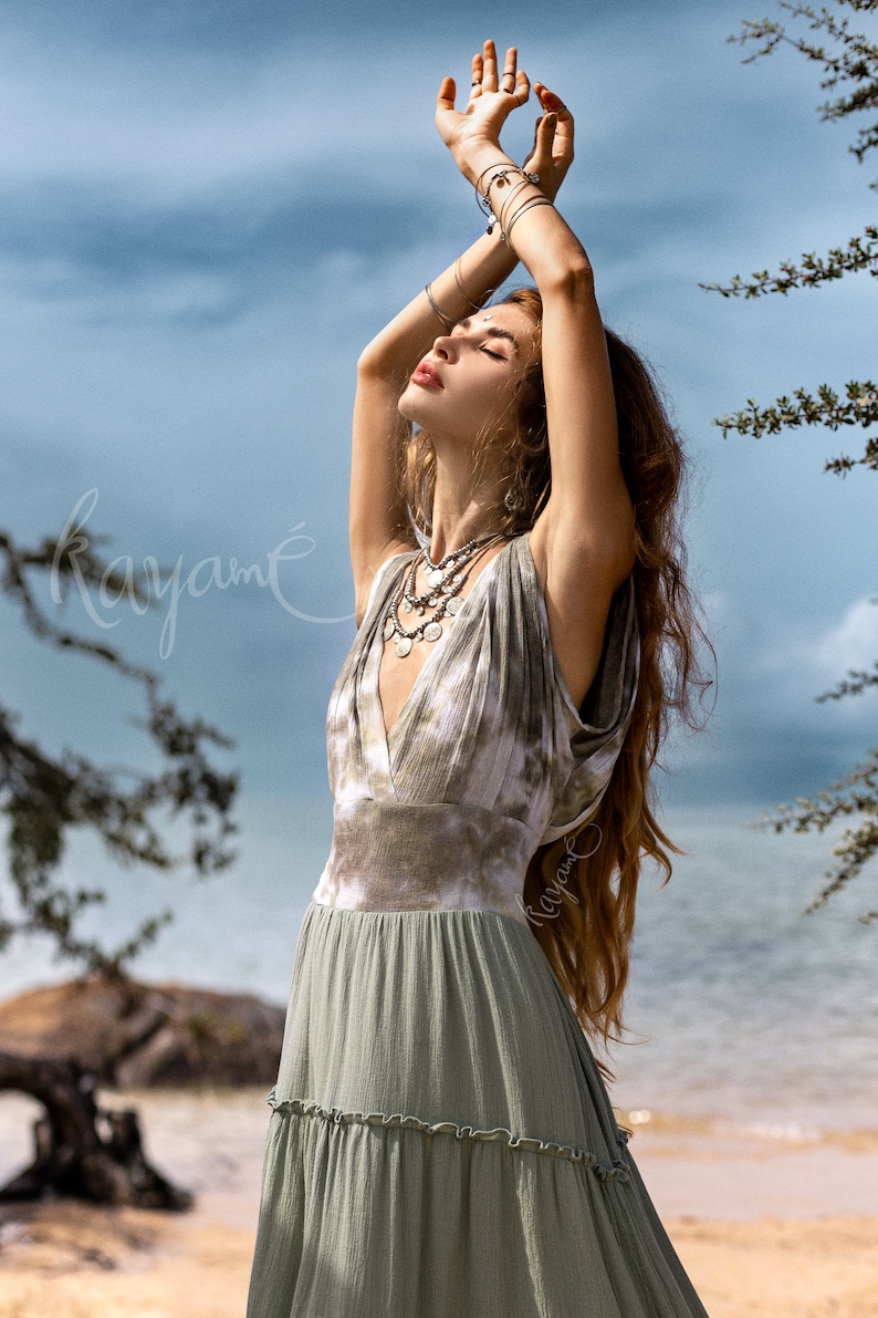 Puede incluir: Una mujer con un vestido maxi verde de tie-dye con falda de capas y un collar plateado. Est&aacute; de pie en una playa con los brazos levantados por encima de la cabeza.