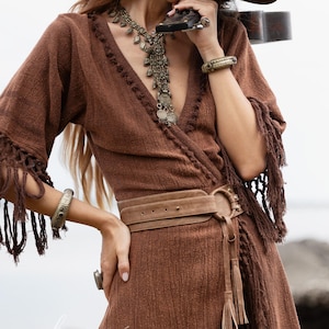 Puede incluir: Un vestido cruzado marr&oacute;n con mangas con flecos y un cintur&oacute;n a juego. El vestido presenta un escote en V y est&aacute; adornado con un collar largo de plata, pulseras y un anillo. El vestido tiene un estilo bohemio.