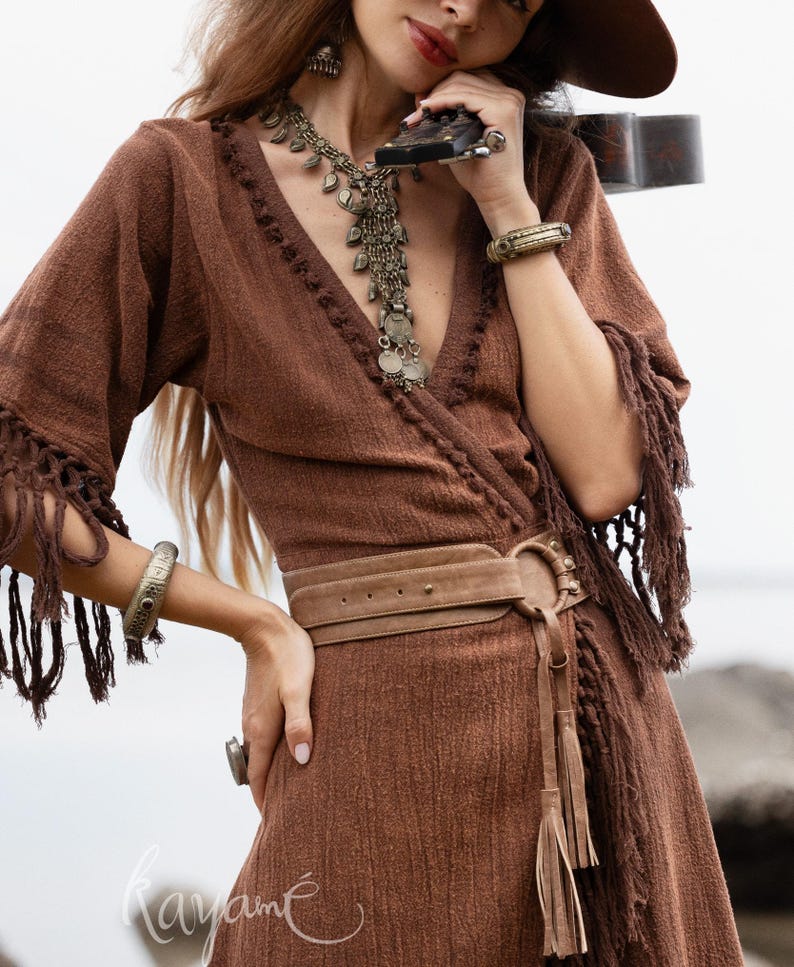 Puede incluir: Un vestido cruzado marr&oacute;n con mangas con flecos y un cintur&oacute;n a juego. El vestido presenta un escote en V y est&aacute; adornado con un collar largo de plata, pulseras y un anillo. El vestido tiene un estilo bohemio.