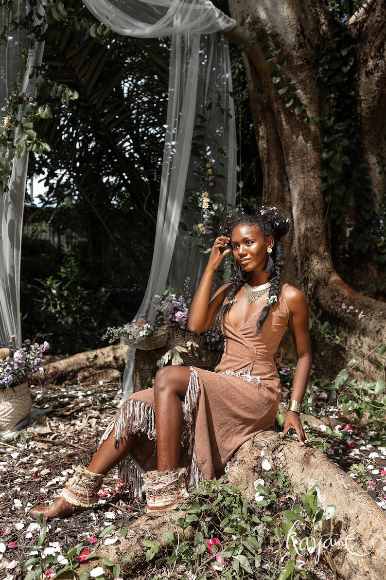 May include: A woman wearing a brown dress with fringe sits on a tree root in a forest setting. She is wearing a gold necklace and earrings. The background is a lush green forest with a white sheer curtain hanging from a tree.