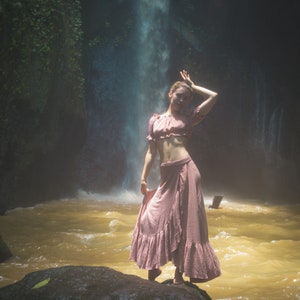 May include: A woman wearing a pink off-the-shoulder top and a long pink skirt stands in front of a waterfall. The water is flowing over rocks and creating a mist.