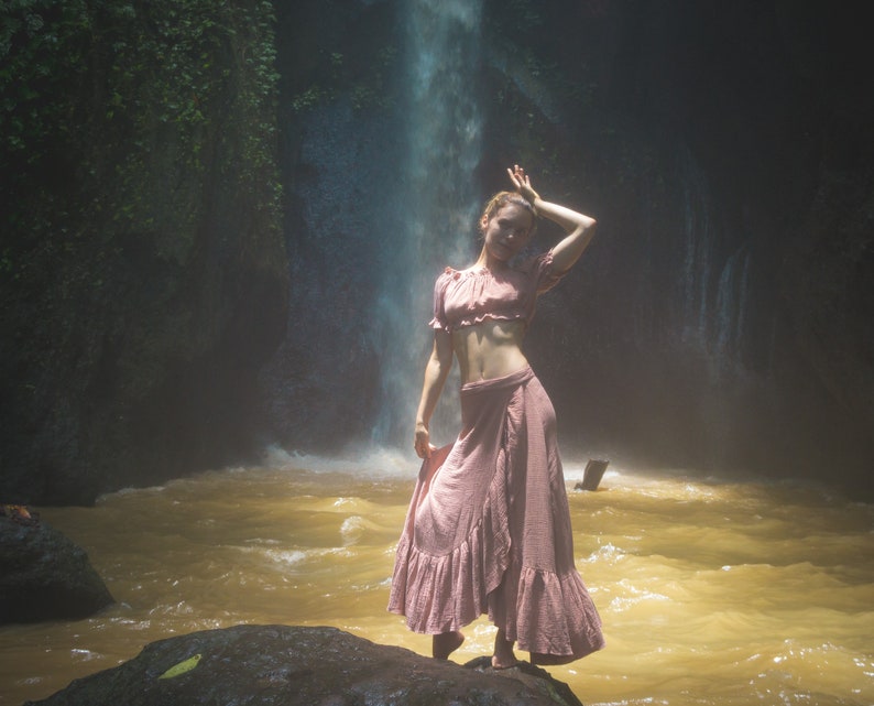 May include: A woman wearing a pink off-the-shoulder top and a long pink skirt stands in front of a waterfall. The water is flowing over rocks and creating a mist.