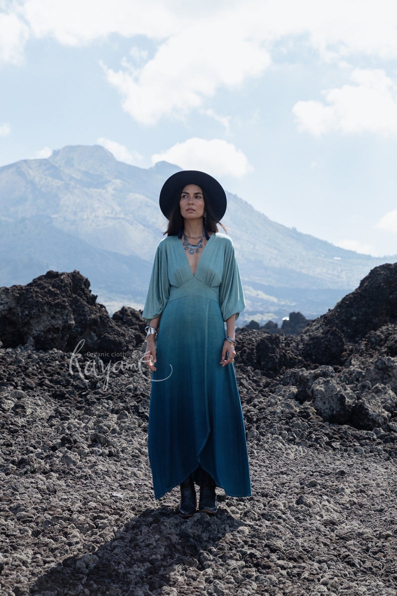 Puede incluir: Un vestido largo hasta el suelo con un escote en V profundo y mangas anchas, que transiciona de un azul verdoso claro en el corpi&ntilde;o a un azul profundo en el dobladillo. La prenda se combina con un sombrero negro de ala ancha, collar y brazaletes.