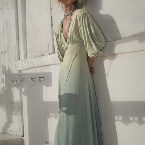 May include: A flowing, light green dress with a V-neck and long, voluminous sleeves. The dress has a subtle ombre effect, fading to a lighter shade at the hem. The model is wearing a matching headscarf and turquoise jewelry. The word "kayant" is written on the wall.