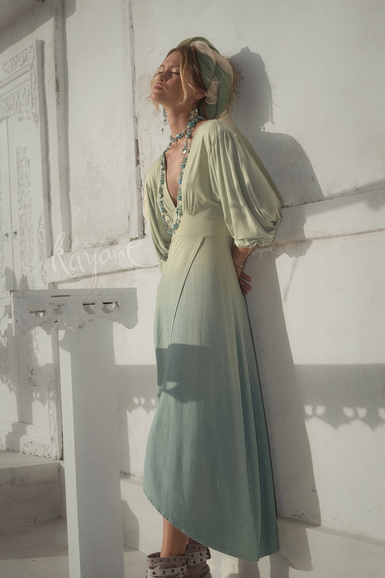 May include: A flowing, light green dress with a V-neck and long, voluminous sleeves. The dress has a subtle ombre effect, fading to a lighter shade at the hem. The model is wearing a matching headscarf and turquoise jewelry. The word "kayant" is written on the wall.
