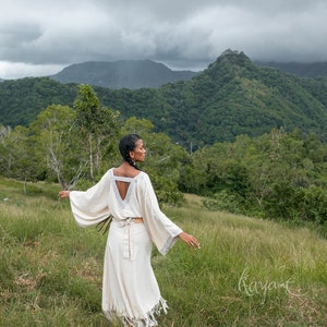 May include: A person wearing a white, flowing dress with a V-neck and a belt, standing in a grassy field with a mountain range in the background.