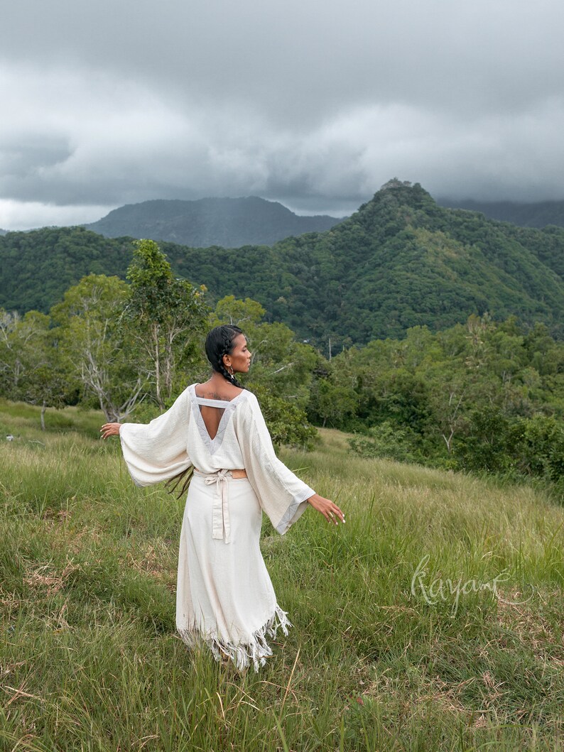 May include: A person wearing a white, flowing dress with a V-neck and a belt, standing in a grassy field with a mountain range in the background.