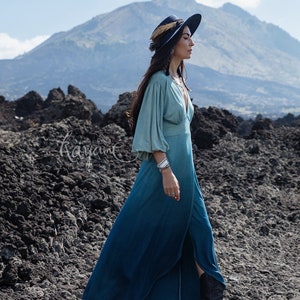 Puede incluir: Un vestido largo hasta el suelo con un escote en V profundo y mangas largas voluminosas, con un degradado de color desde el verde azulado claro en la parte superior hasta el azul oscuro en el dobladillo. Incluye una abertura frontal y el texto Kayame.
