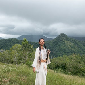 May include: A woman wearing a white, flowing dress with a brown belt and long, dark braids stands in a grassy field with a mountain range in the background. The sky is overcast with clouds.