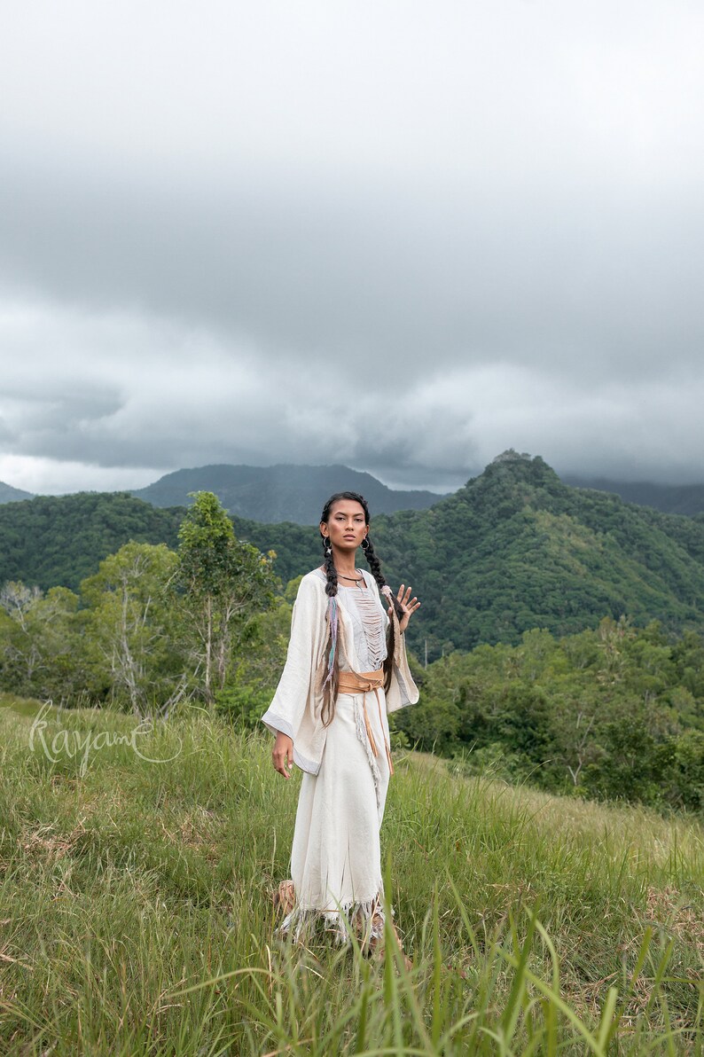 May include: A woman wearing a white, flowing dress with a brown belt and long, dark braids stands in a grassy field with a mountain range in the background. The sky is overcast with clouds.