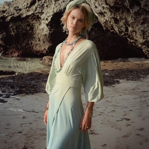 May include: A flowing, light green dress with a deep V-neck and long sleeves. The dress is paired with a matching headscarf and turquoise jewelry. The image is set on a sandy beach with a large rock formation in the background.