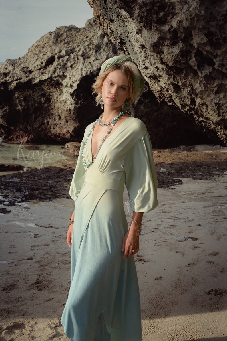 May include: A flowing, light green dress with a deep V-neck and long sleeves. The dress is paired with a matching headscarf and turquoise jewelry. The image is set on a sandy beach with a large rock formation in the background.