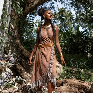 May include: A woman wearing a brown wrap dress with fringe details and a gold necklace stands in a forest setting. She is wearing brown boots with fringe details and has her hair in braids. The background is a lush forest with green trees and foliage.