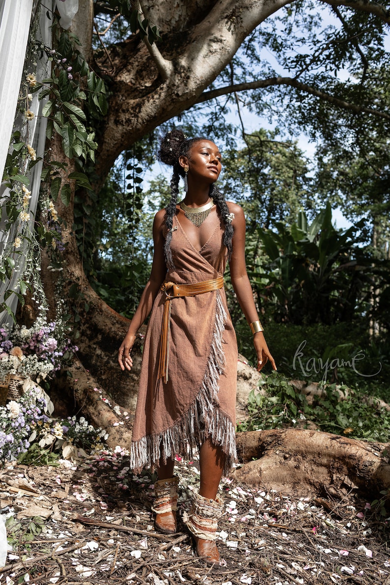 May include: A woman wearing a brown wrap dress with fringe details and a gold necklace stands in a forest setting. She is wearing brown boots with fringe details and has her hair in braids. The background is a lush forest with green trees and foliage.