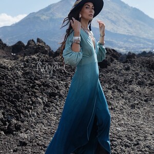 Puede incluir: Un vestido largo de manga larga con un degradado de color que va del verde salvia claro en el corpi&ntilde;o al azul cerceta profundo en el dobladillo, con escote en V profundo, mangas fruncidas y falda fluida, acompa&ntilde;ado de un sombrero y botas.
