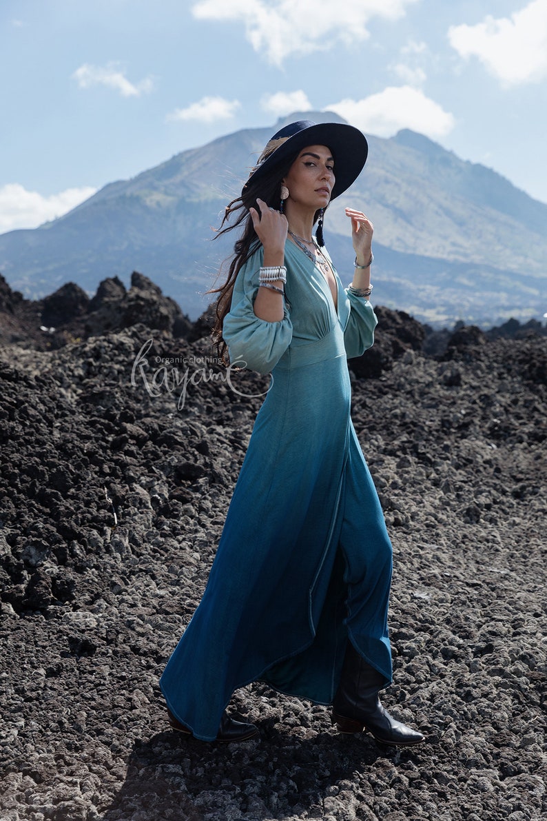 Puede incluir: Un vestido largo de manga larga con un degradado de color que va del verde salvia claro en el corpi&ntilde;o al azul cerceta profundo en el dobladillo, con escote en V profundo, mangas fruncidas y falda fluida, acompa&ntilde;ado de un sombrero y botas.